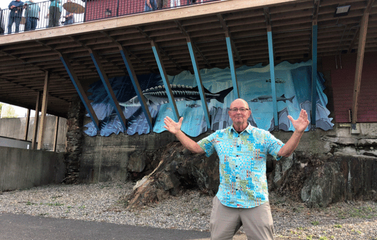 Artist David Hurley poses near his sturgeon mural on the Bayview Events Center, near Belfast’s harbor walk. PHOTO: TOM GROENING